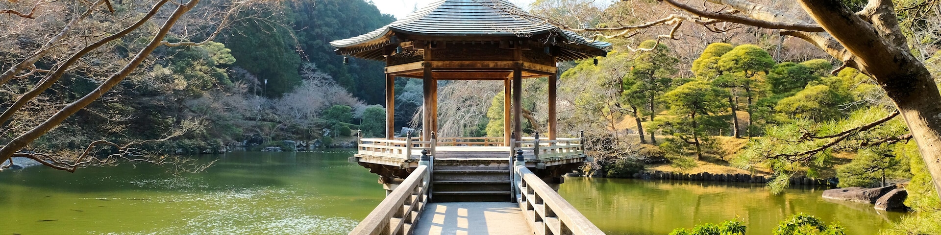 A traditional Japanese pavilion over a bridge leading to a calm lake surrounded by trees.