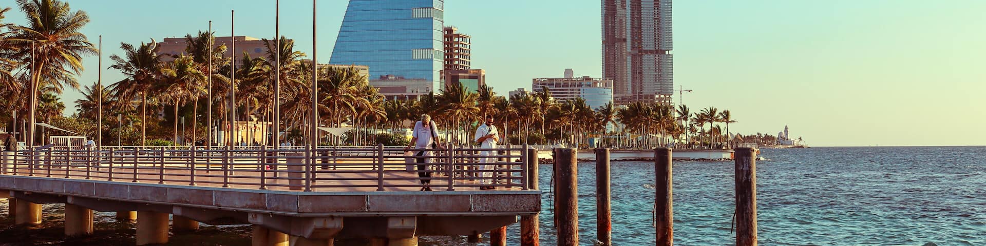 A waterfront promenade with palm trees, a modern glass building, and a construction site.