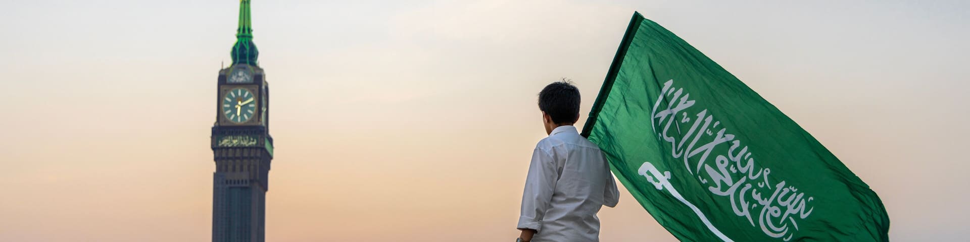 A person holding a flag with Arabic script, standing in front of a tall building with a clock and a spire, with a cityscape in the background.