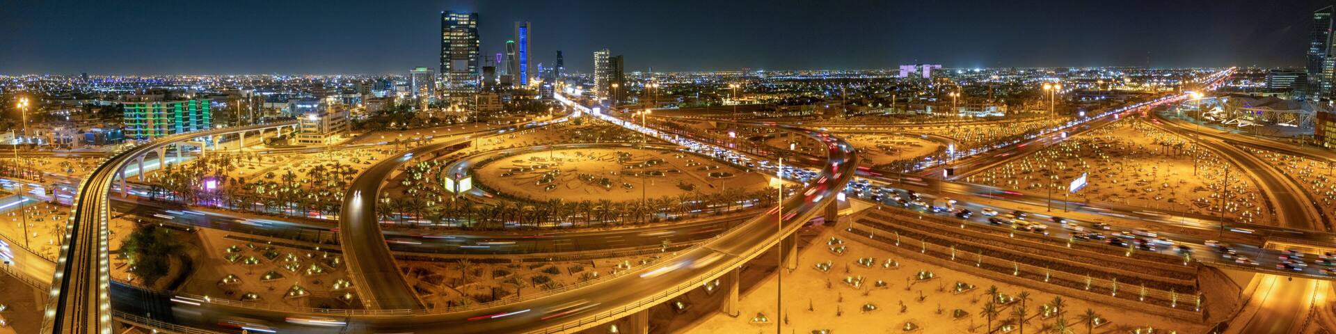 A nighttime aerial view of a complex highway interchange with multiple overpasses and a cityscape in the background.