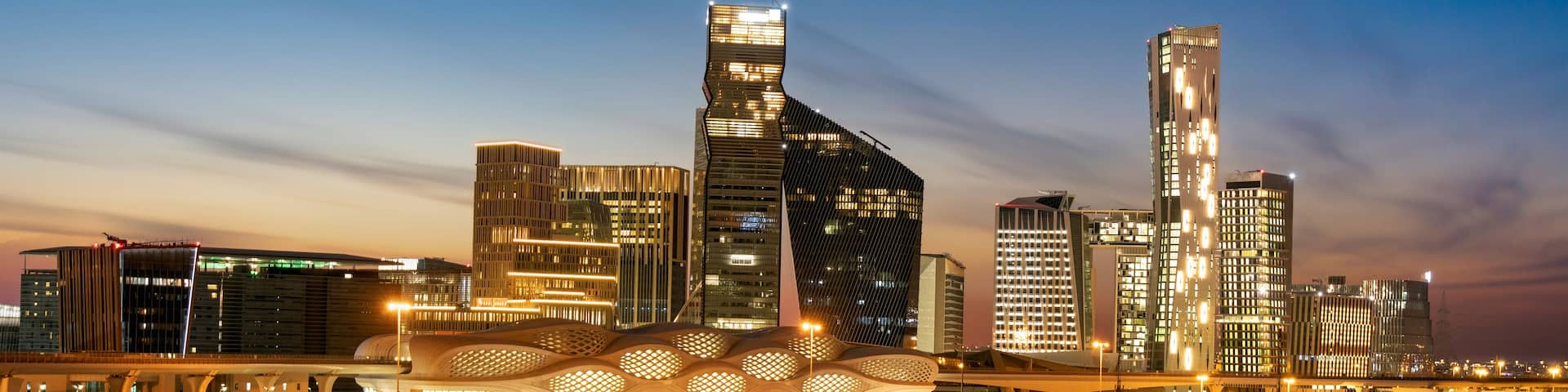 A city skyline at dusk with illuminated buildings and a distinctive modern architectural structure.