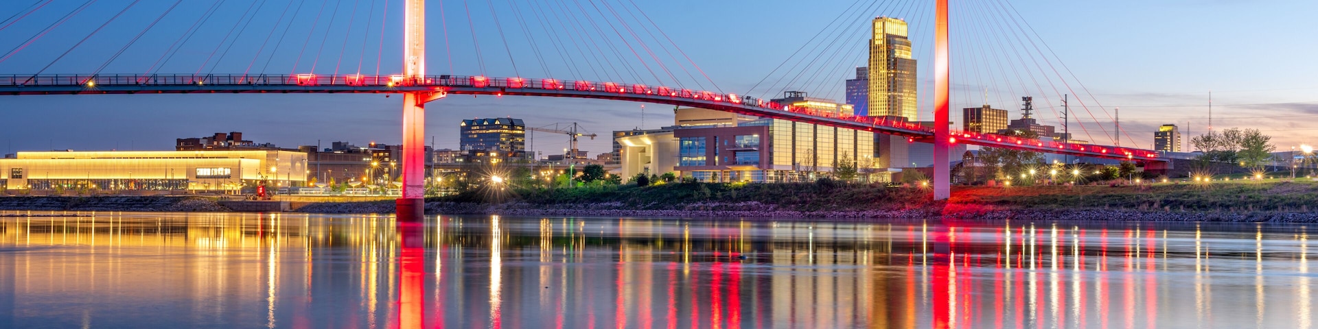 Un puente con luces rojas reflejadas en el agua, un horizonte urbano al fondo y un cielo despejado.