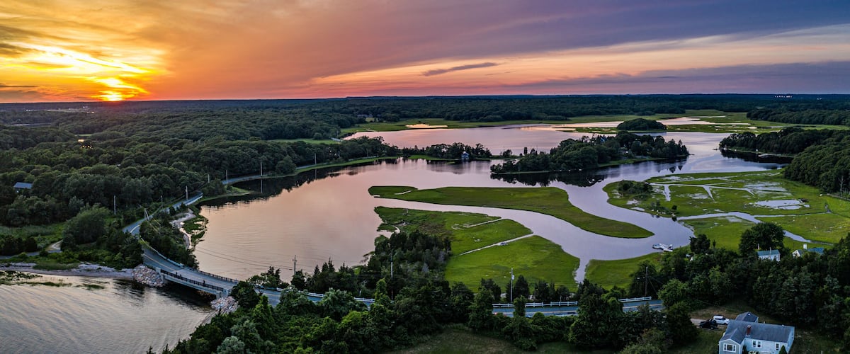 A sunset over a river with a bridge, surrounded by greenery and a small house.