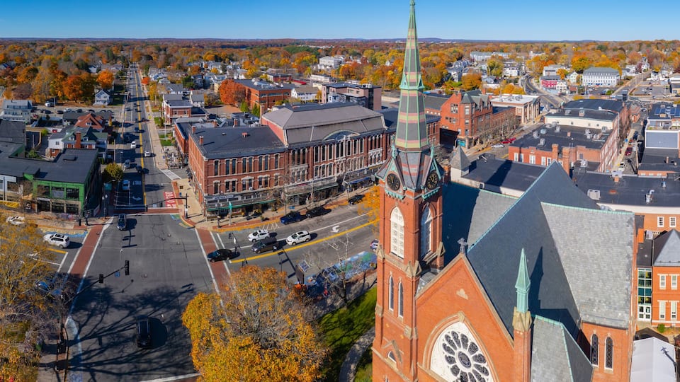 An aerial view of a town with a prominent church steeple, a main street, and autumn-colored trees.