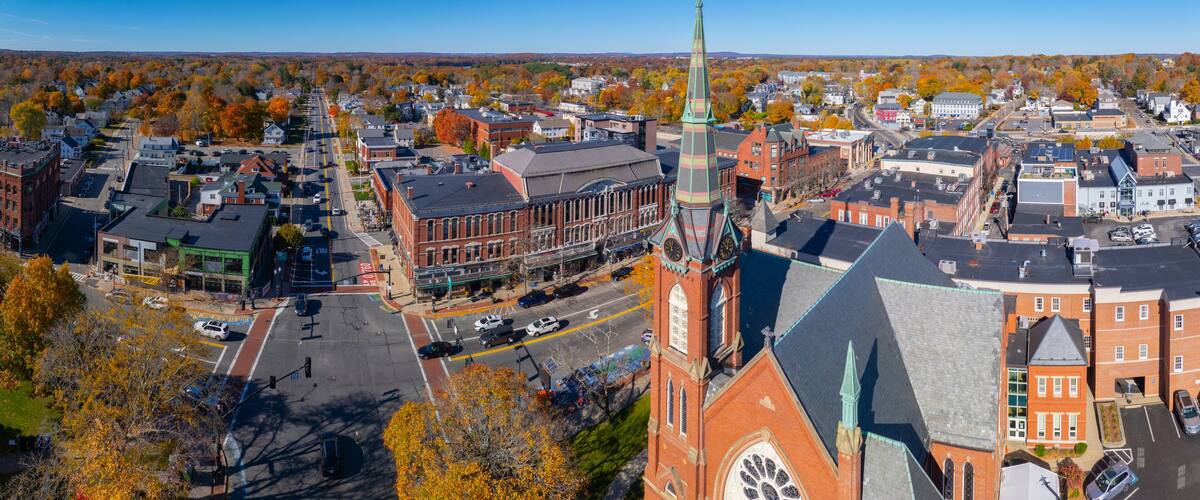 An aerial view of a town with a prominent church steeple, a main street, and autumn-colored trees.