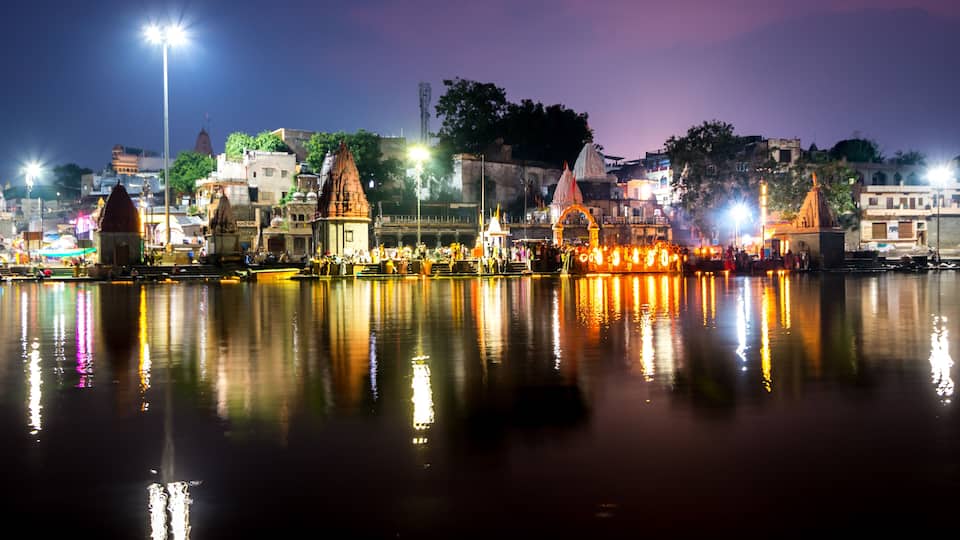 A nighttime view of a riverside with illuminated buildings and a bridge reflecting on the water.