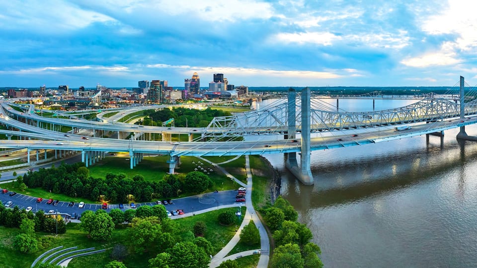 A multi-level highway interchange with a large suspension bridge over a river, surrounded by urban buildings and greenery.