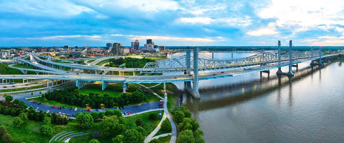 A multi-level highway interchange with a large suspension bridge over a river, surrounded by urban buildings and greenery.