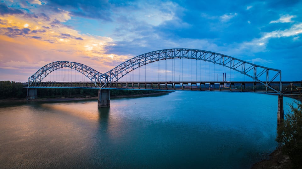 A steel arch bridge over a calm river at sunset.