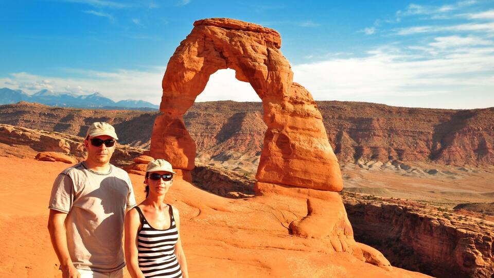 A couple standing in front of a natural rock arch formation with a desert landscape in the background.