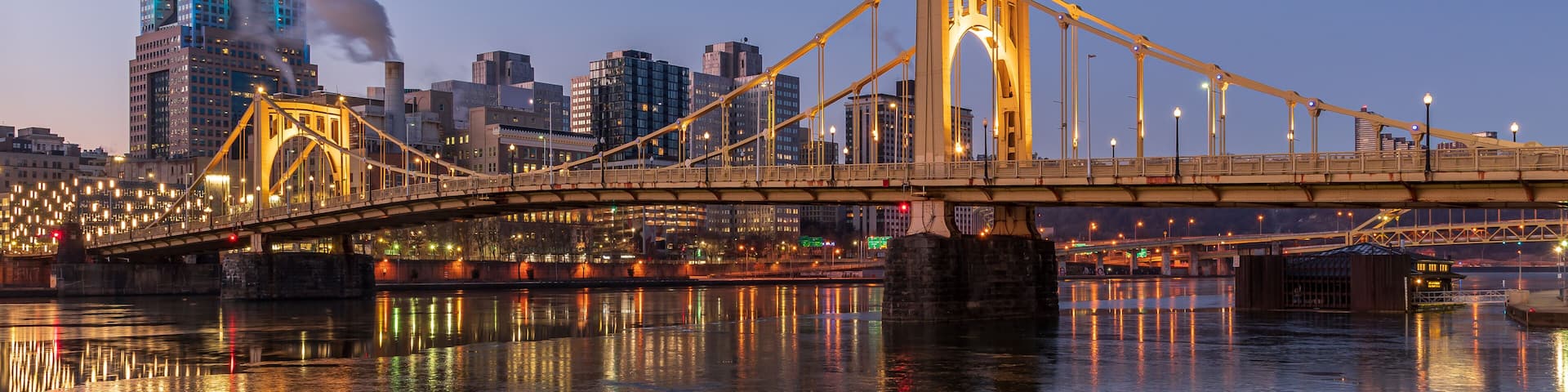 A bridge with illuminated lights reflecting on the water, a city skyline in the background, and a smokestack emitting smoke.