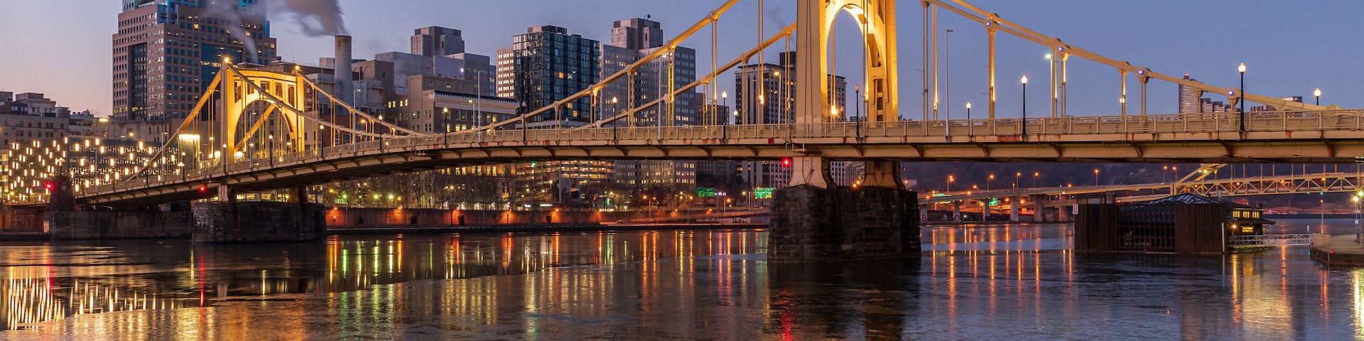 A bridge with illuminated lights reflecting on the water, a city skyline in the background, and a smokestack emitting smoke.