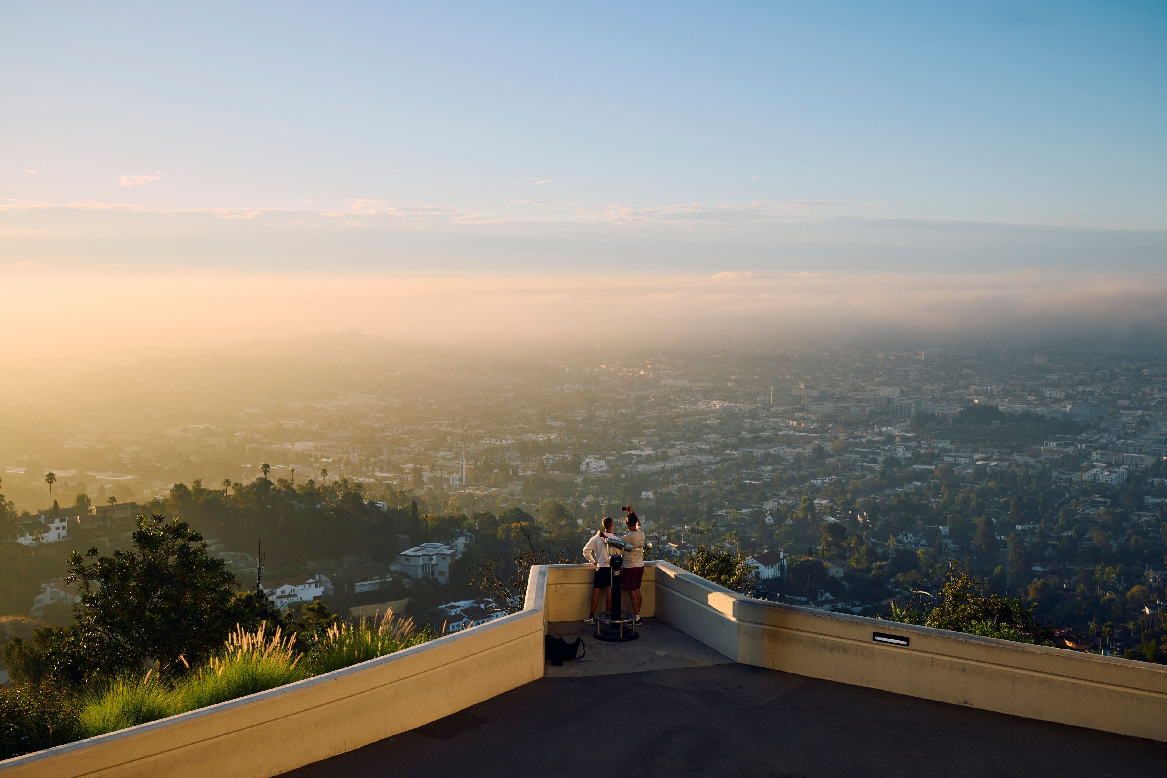 A panoramic view of a cityscape from a high vantage point, featuring a clear sky, distant mountains, and a dense urban area below.