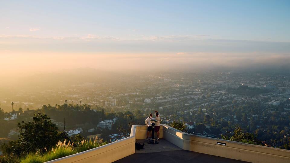 A panoramic view of a cityscape from a high vantage point, featuring a clear sky, distant mountains, and a dense urban area below.