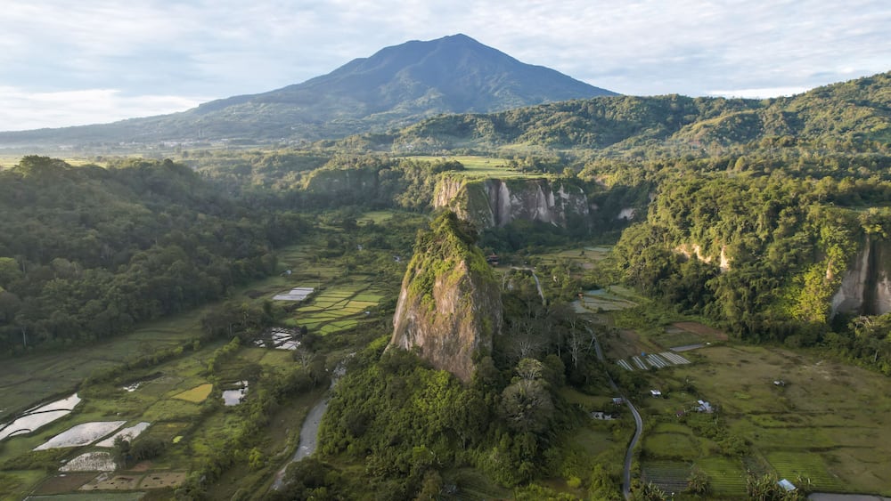 A mountainous landscape with terraced fields, a prominent rocky peak, and a distant volcano.