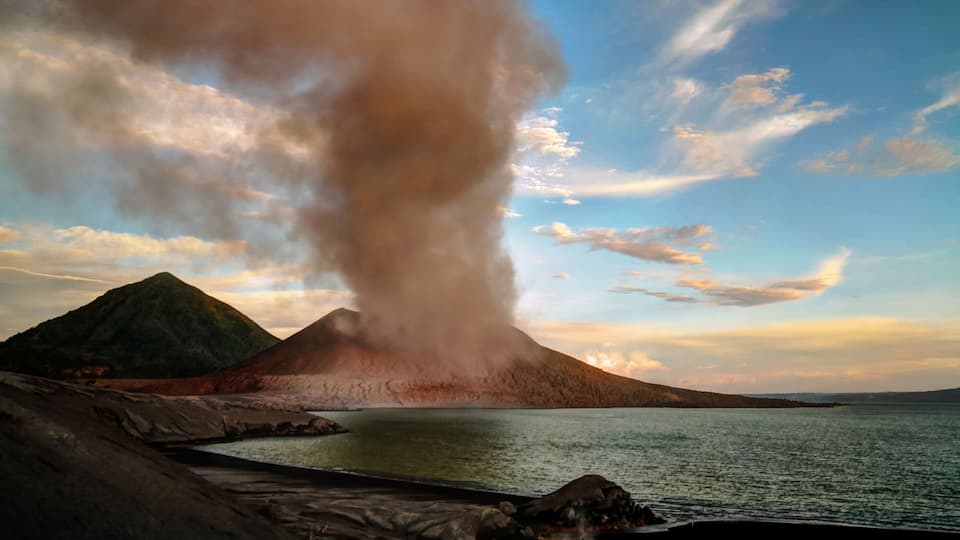 A volcano with a plume of smoke, a body of water, and a mountainous landscape.