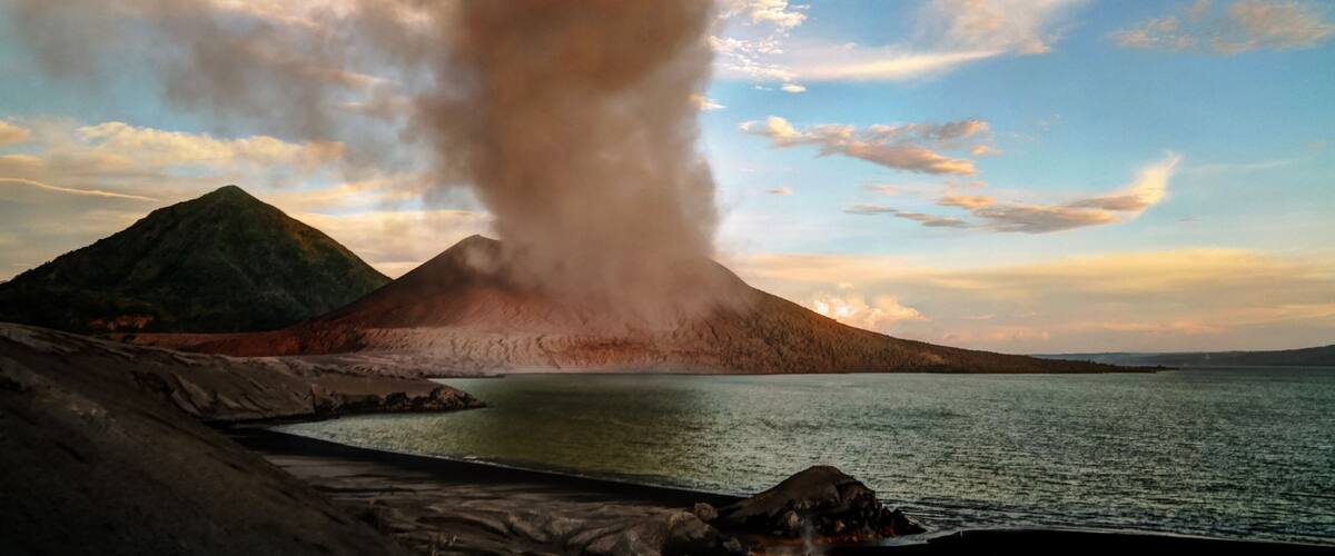 A volcano with a plume of smoke, a body of water, and a mountainous landscape.