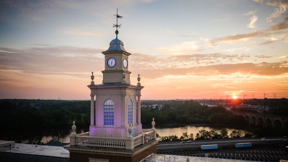 A clock tower with a weather vane, a river, and a bridge in the background during sunset.