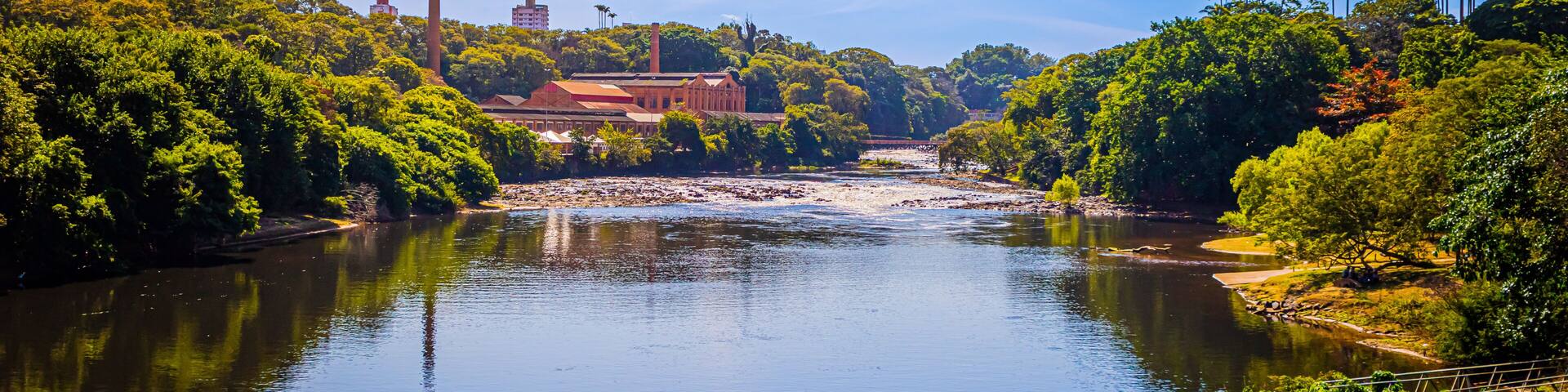 A river with a bridge, surrounded by dense greenery and industrial buildings in the background.
