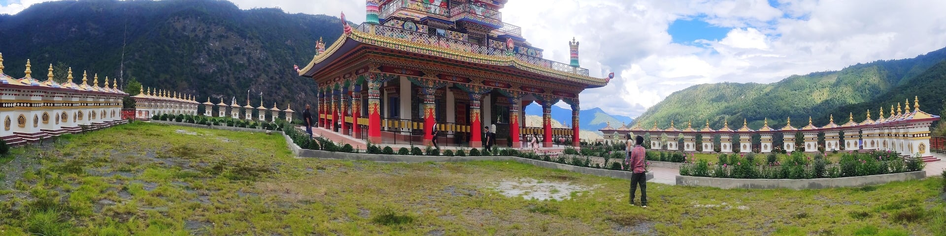 A large golden statue atop a colorful temple complex surrounded by lush green mountains.