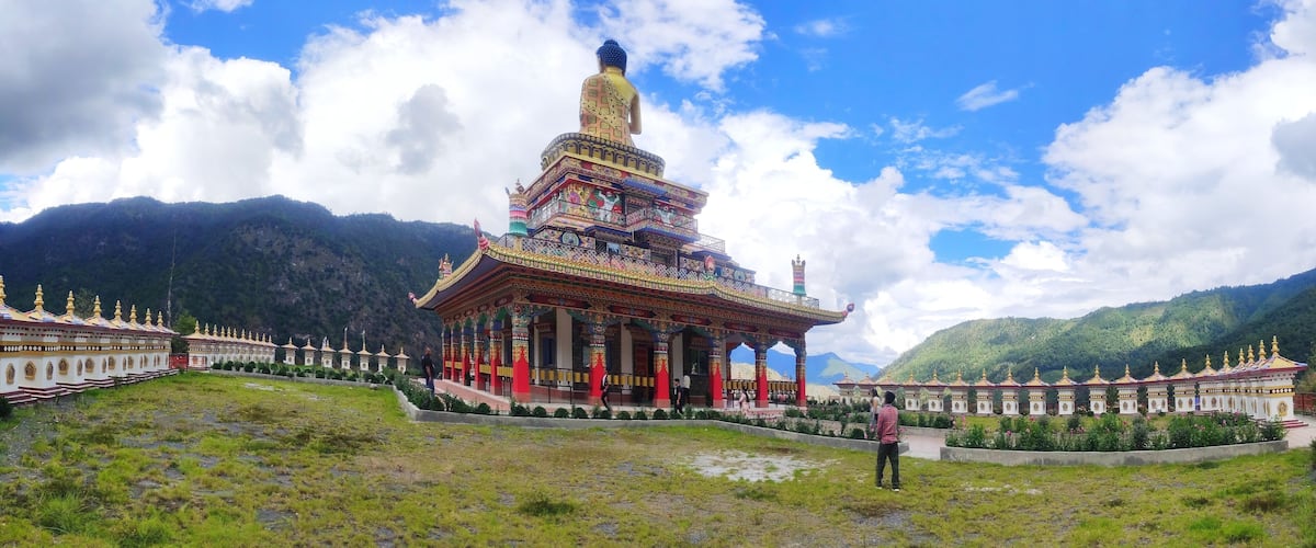 A large golden statue atop a colorful temple complex surrounded by lush green mountains.
