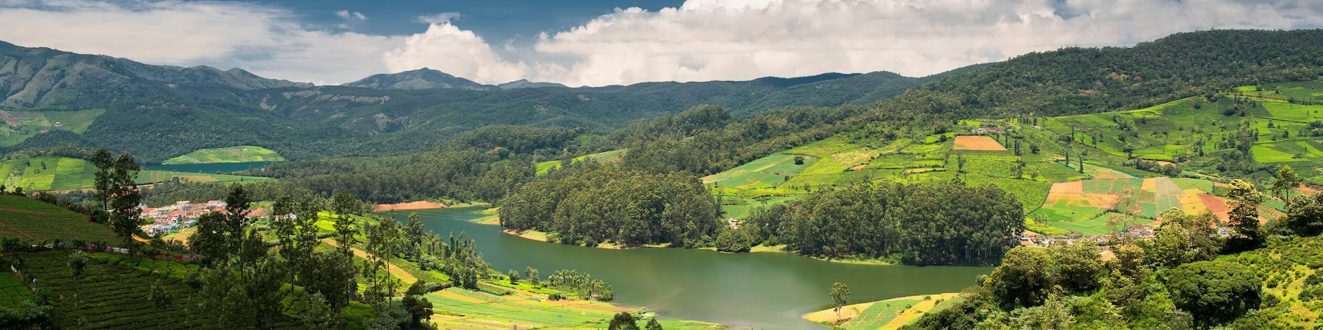 A lush valley with terraced tea plantations, a river, and rolling hills under a partly cloudy sky.