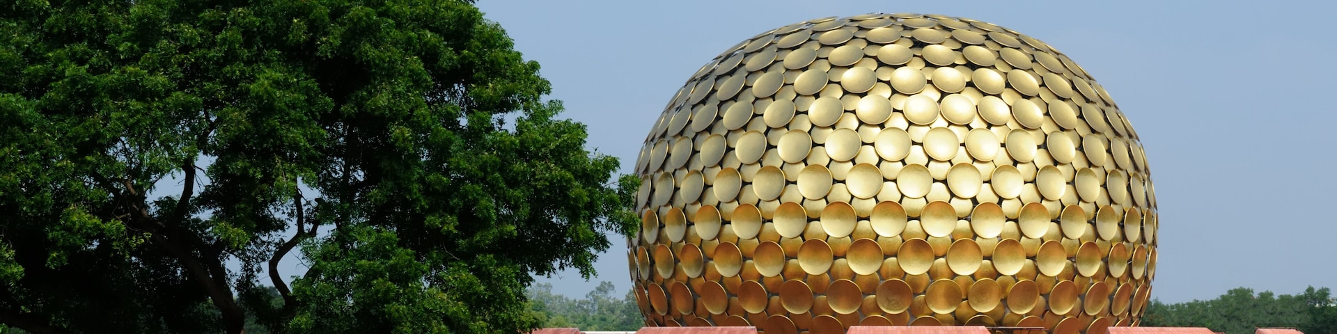 A large golden sphere with a pattern of circular panels, surrounded by red pyramids and a green field with a tree.