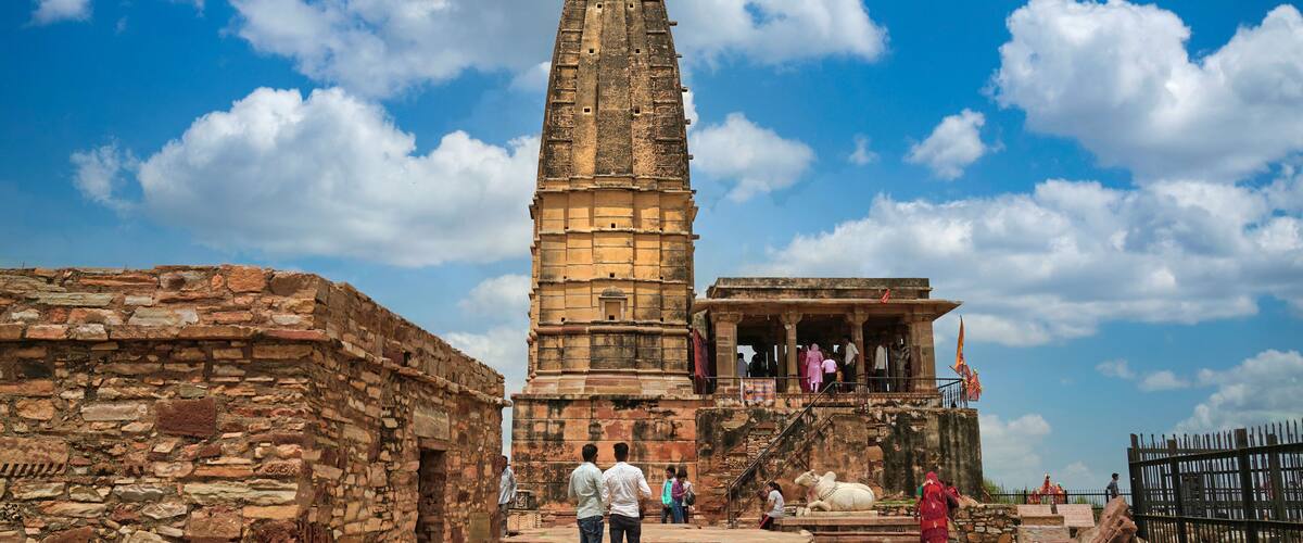An ancient stone structure with a tall spire, a stone wall, and a small pavilion with people around it.