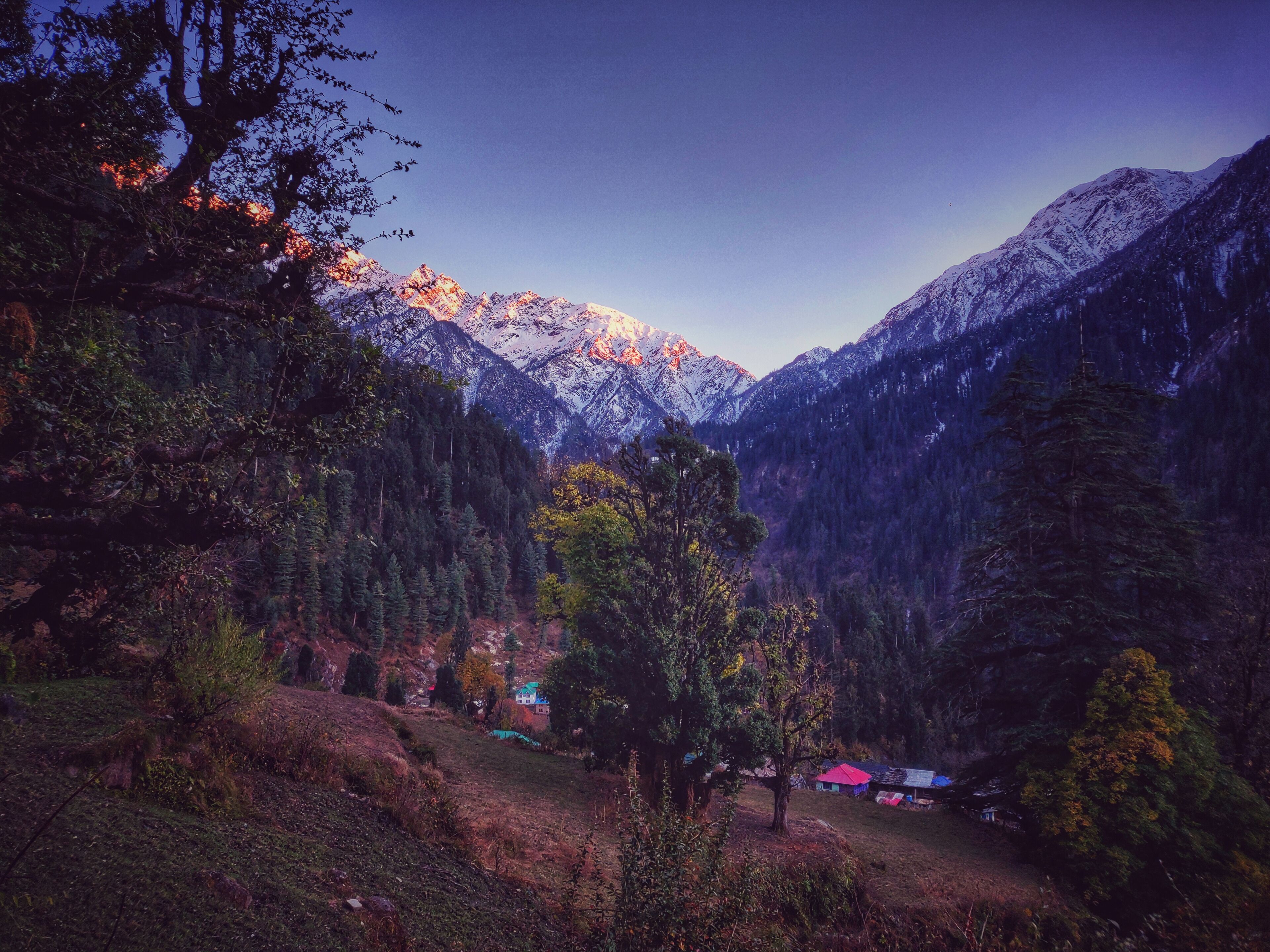 A mountainous landscape with snow-capped peaks, dense forests, and a small settlement with colorful buildings.