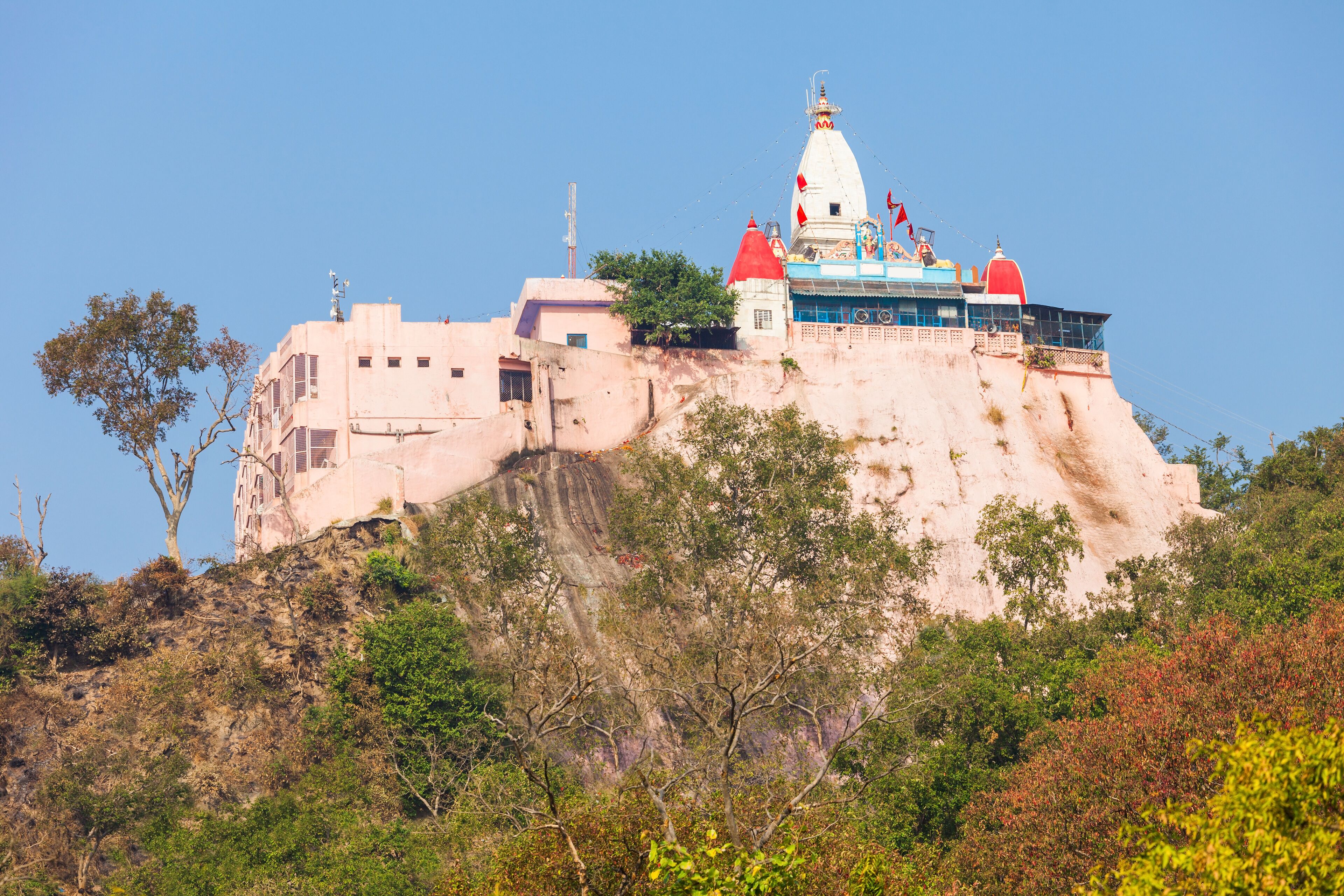 A large, multi-story building with a red dome and a white spire on a hill, surrounded by trees and rocky terrain.