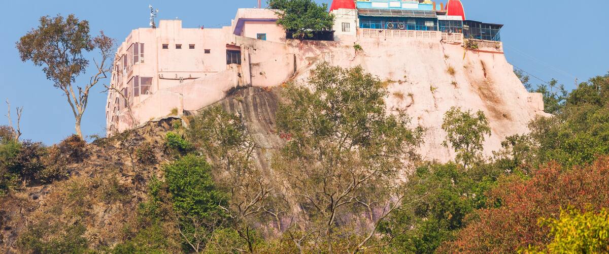 A large, multi-story building with a red dome and a white spire on a hill, surrounded by trees and rocky terrain.