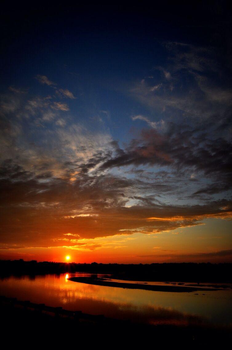 Un coucher de soleil sur un lac paisible, avec la silhouette d’arbres et un ciel dégagé.