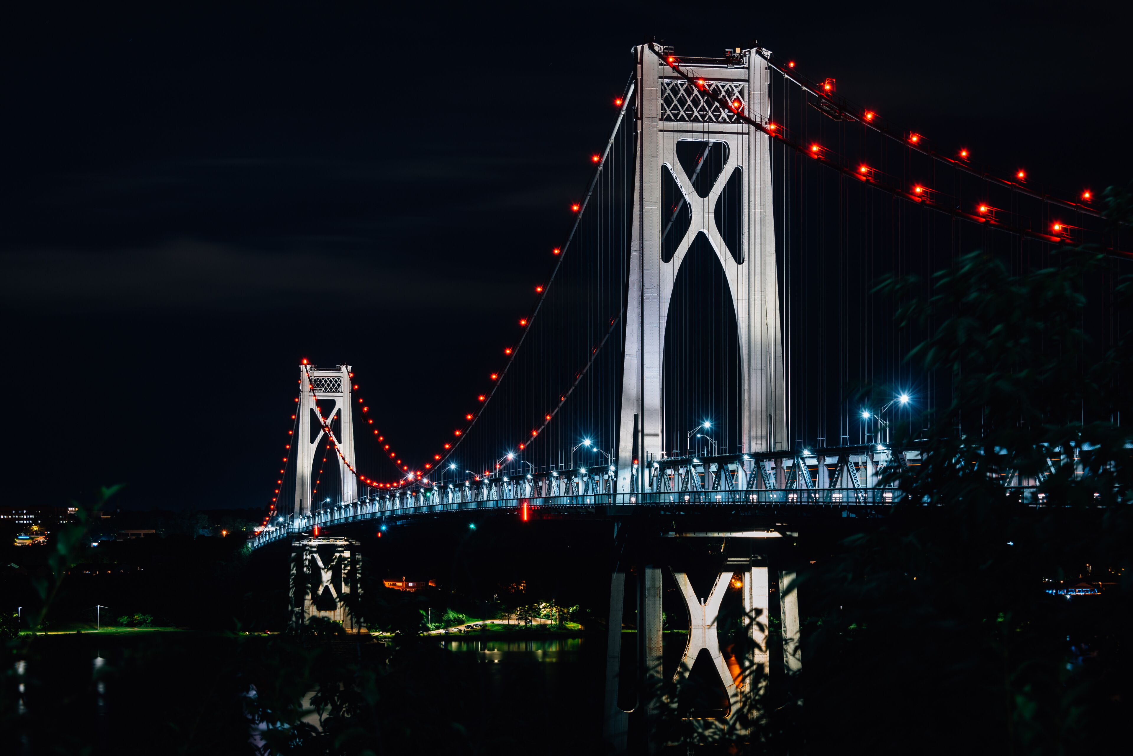 A nighttime view of a large suspension bridge with red lights, featuring two towers and a central arch.