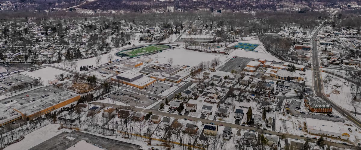 A snowy urban landscape with a large sports field, residential areas, and a distant horizon.