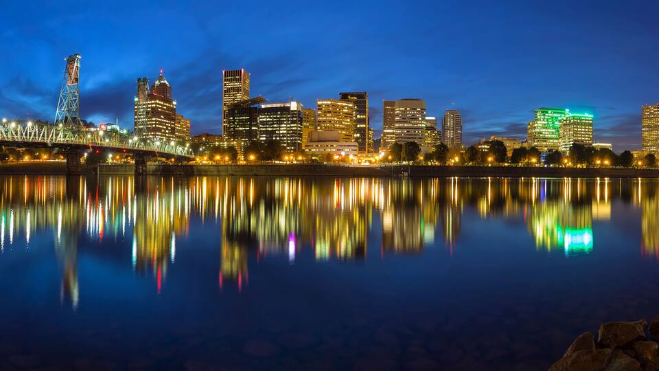 A nighttime city skyline reflected in a calm body of water, featuring illuminated buildings and a bridge.