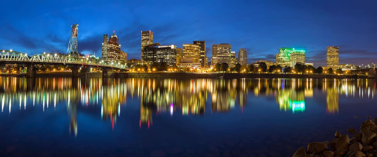 A nighttime city skyline reflected in a calm body of water, featuring illuminated buildings and a bridge.