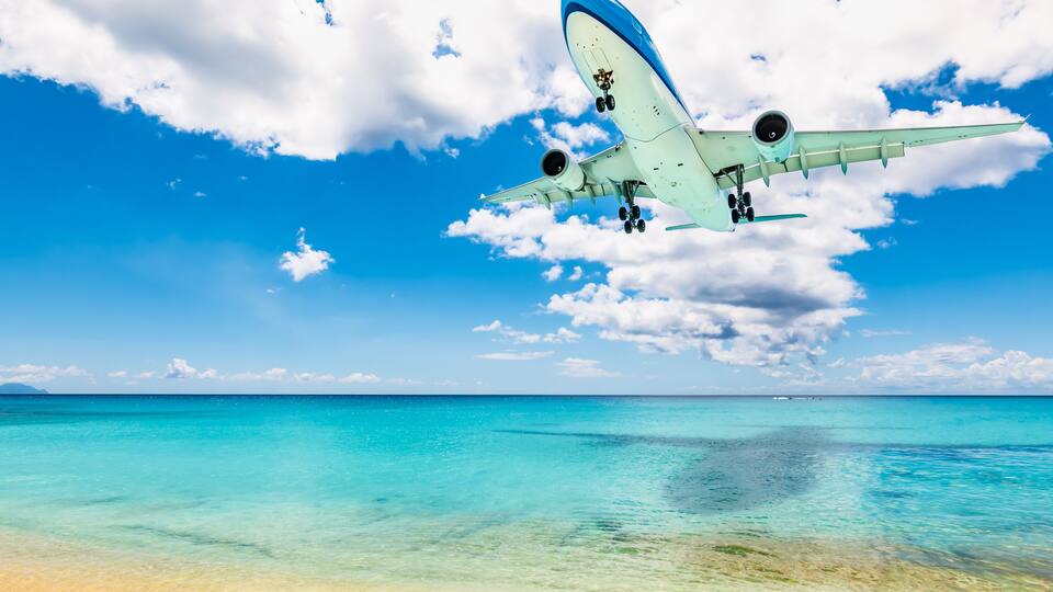 A clear blue sky, turquoise sea, and a plane flying over a beach.