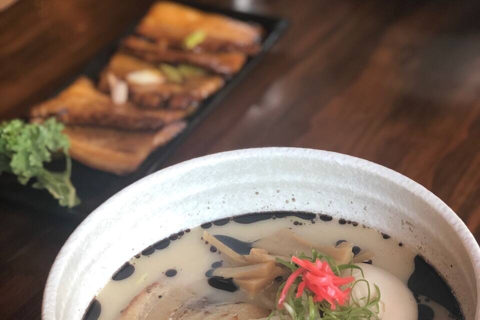 A bowl of soup with noodles and vegetables, a metal cup, and a plate of food in the background.