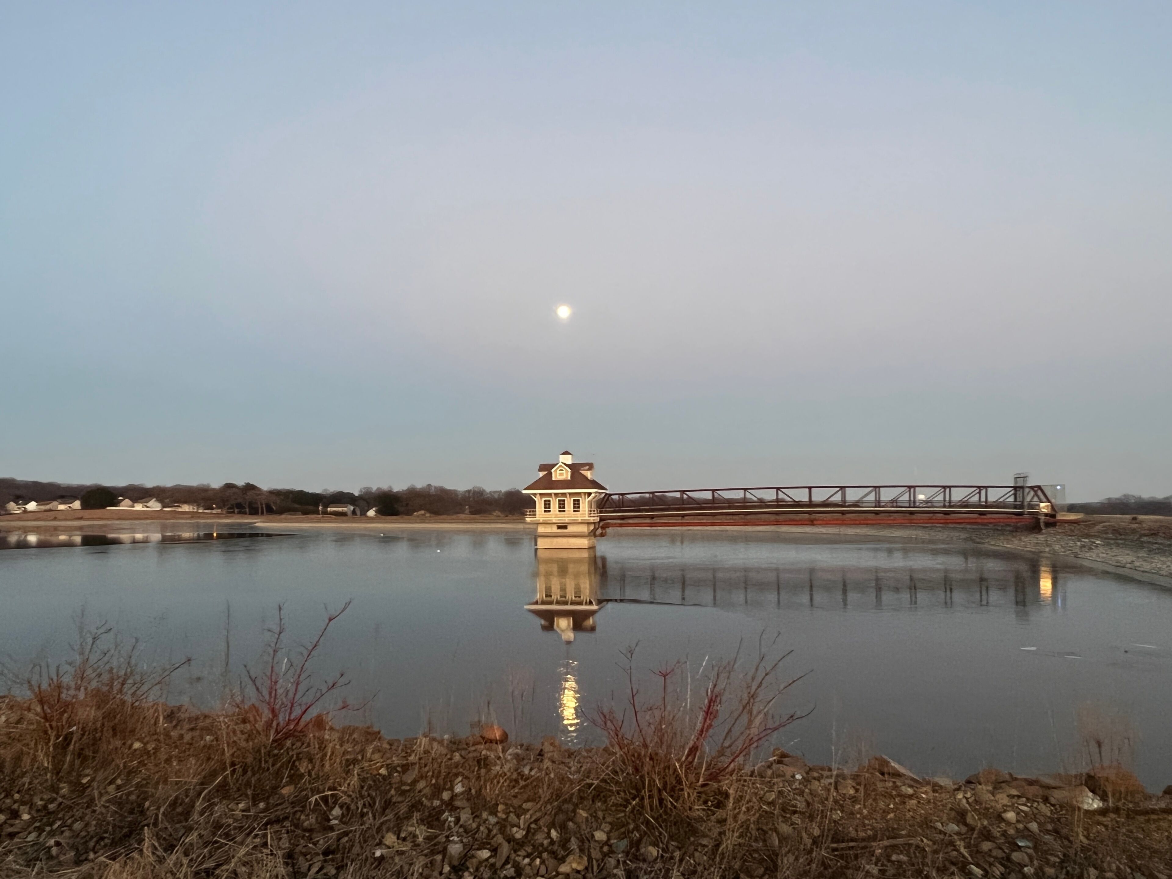 A calm lake with a small tower-like structure in the center, a bridge in the background, and a clear sky.