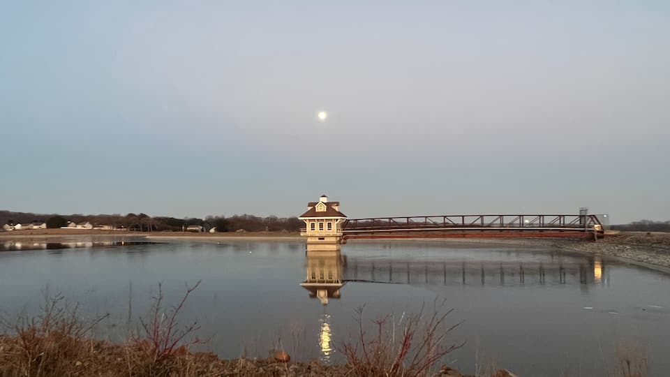 A calm lake with a small tower-like structure in the center, a bridge in the background, and a clear sky.