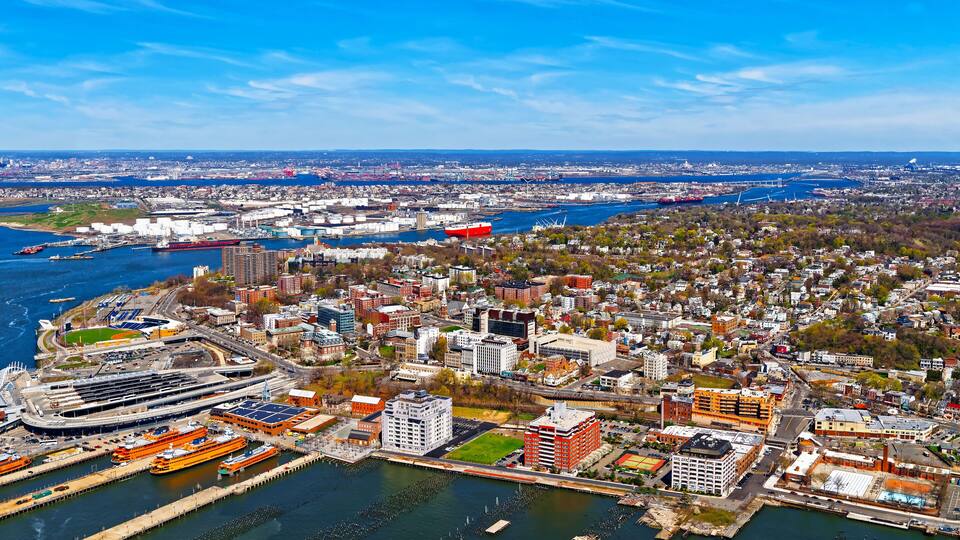 A coastal cityscape with a prominent harbor, including a large cruise ship docked at the pier, and a dense urban area with various buildings and greenery.
