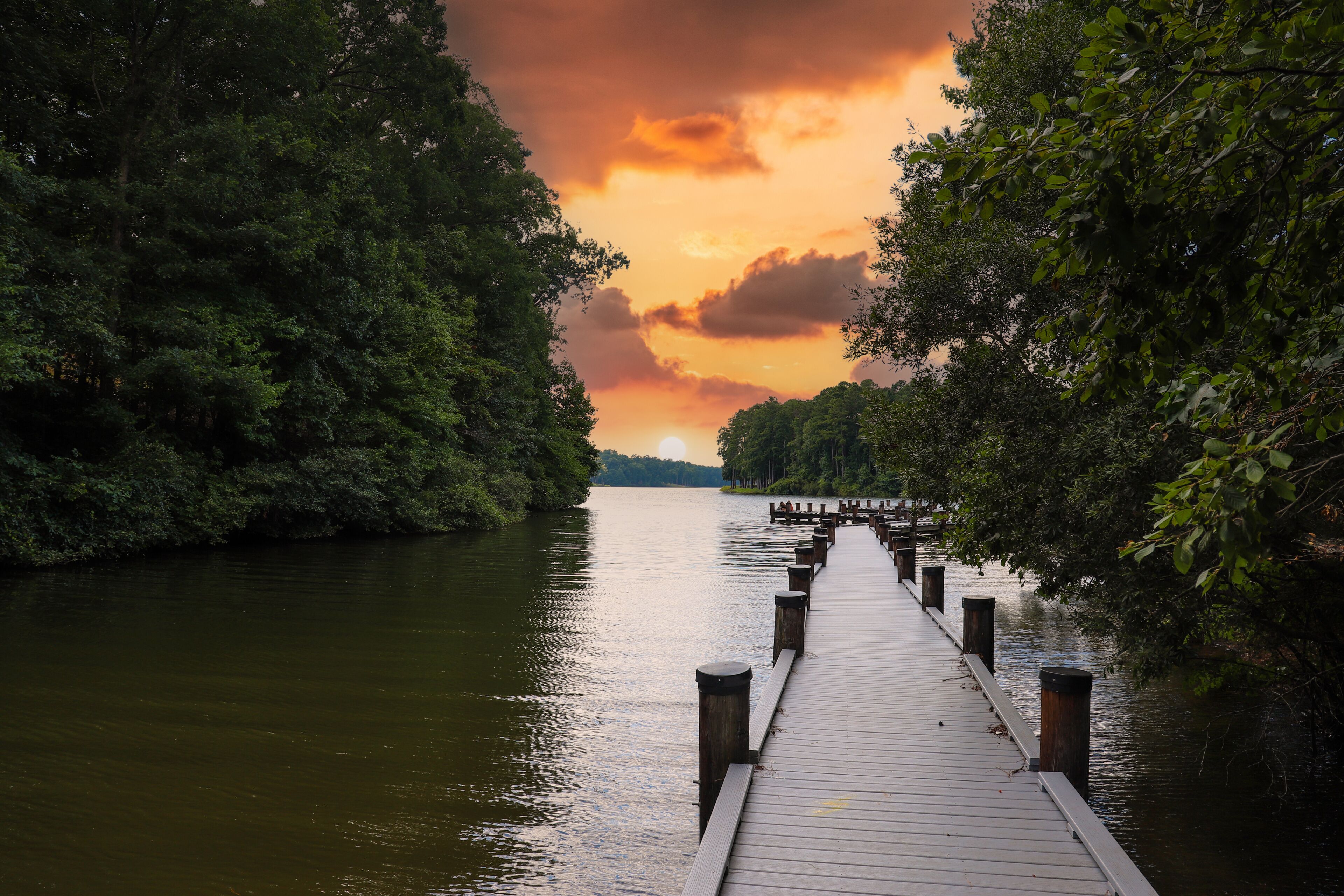 a gorgeous shot of the still blue lake water with a wooden dock over the water surrounded by lush green trees with powerful clouds at sunset at Lake Kedron in Peachtree City Georgia USA