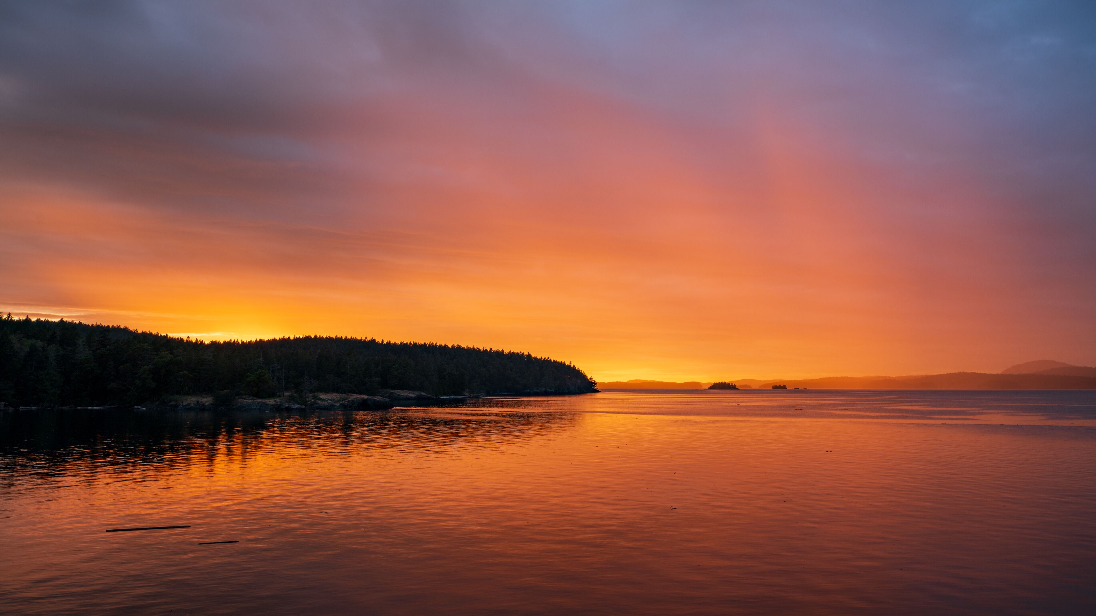 A sunset over a calm lake with a forested island and distant mountains.