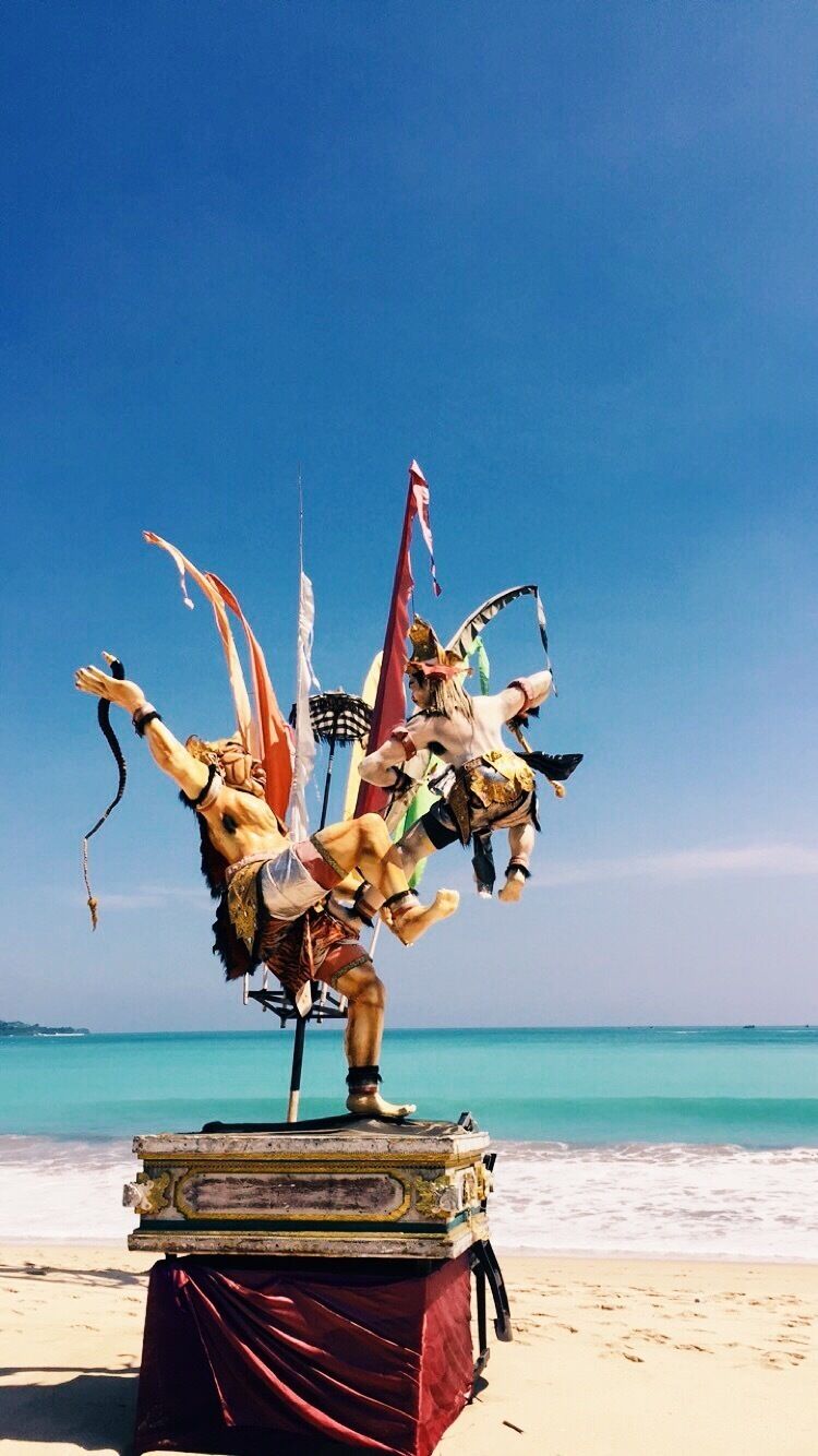 A statue of a figure with multiple arms on a pedestal by the beach.