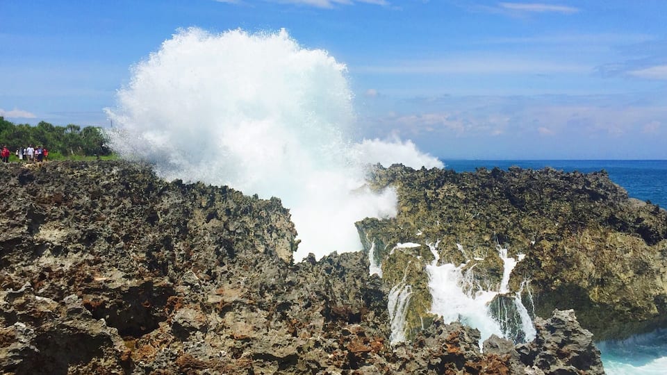 Une côte rocheuse où une grosse vague s’écrase contre les rochers, créant un effet de cascade.