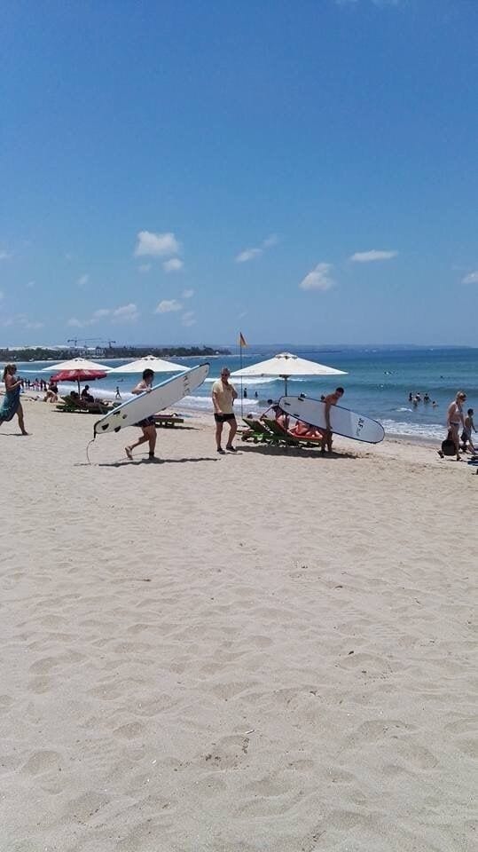 A beach with people walking, some carrying surfboards, and umbrellas set up on the sand.