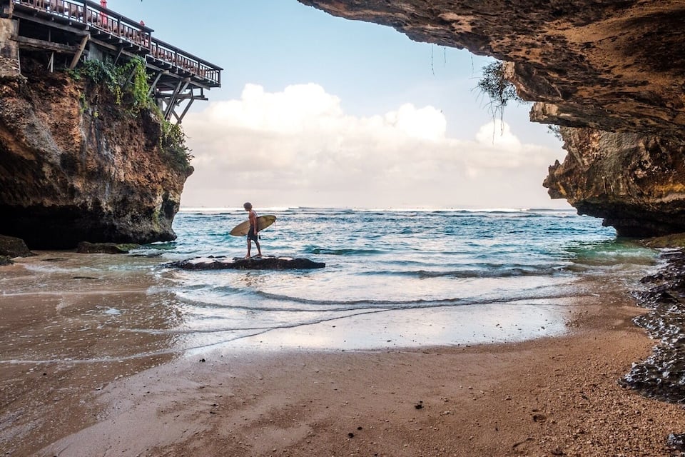 Uma pessoa em uma rocha com uma prancha de surf, observando uma praia com penhascos rochosos e uma ponte.