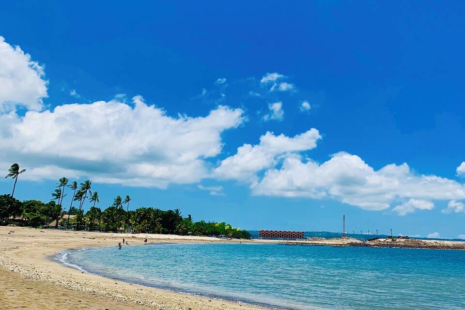 A beach with clear blue water, palm trees, and a distant building.