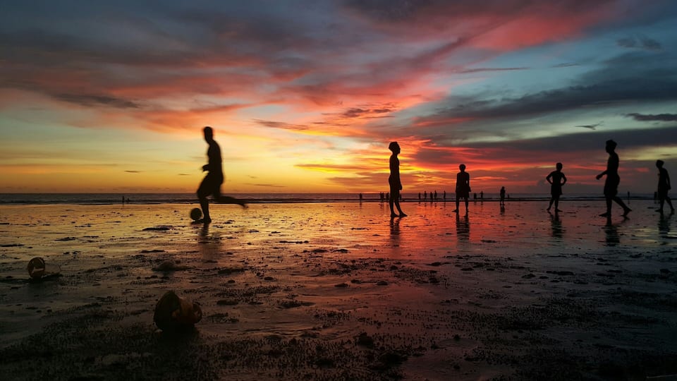 A beach at sunset with people playing soccer.
