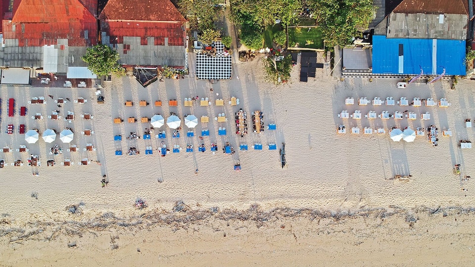 An aerial view of a beach with rows of sunbeds and umbrellas, surrounded by buildings with red and blue facades.